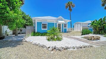 A two-story house with a blue exterior and a white roof, surrounded by a gravel driveway and tropical vegetation.