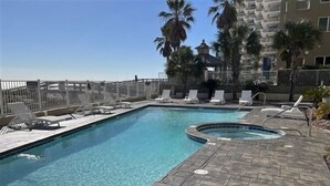 Una piscina con sillones de descanso y un jacuzzi, rodeada de palmeras y un edificio.