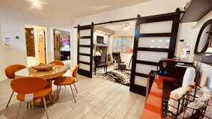 A modern dining area with a round table and orange chairs, a black-framed sliding door, and a kitchen in the background. - Loft Escape Artistic (Surprise)