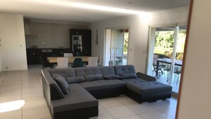 Uma sala de estar moderna com um sofá seccional, uma área de refeições com cadeiras e uma cozinha com eletrodomésticos.