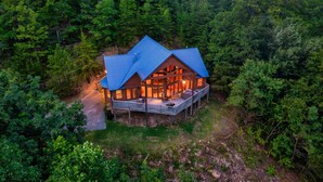 A cabin with a blue roof, surrounded by dense forest. It features a deck with outdoor seating and a visible interior with warm lighting. - Private cabin with stunning views near Pigeon Forge (Sevierville)