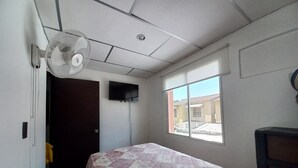 Un dormitorio con cama, ventilador de techo, televisor montado en la pared y ventana con vistas a edificios.