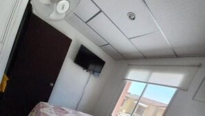 Un dormitorio con una cama cubierta por una colcha estampada con motivos florales, un ventilador de techo, un televisor montado en la pared y una ventana que ofrece vistas al exterior.