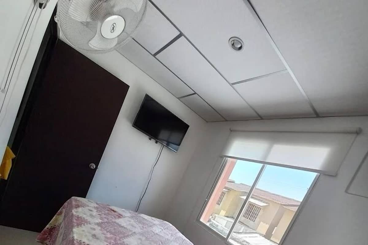 A bedroom with a bed covered in a floral-patterned bedspread, a ceiling fan, a TV mounted on the wall, and a window providing a view of the outside.