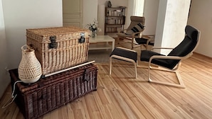 A living room with a large wicker chest, a wicker basket, a black cushioned chair, a small table, and a bookshelf in the background. - Bed and breakfast / gîte with SPA (Massages on reservation) (Domessargues)