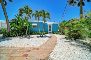 A blue house with a paved pathway leading to the entrance, surrounded by palm trees and tropical plants.