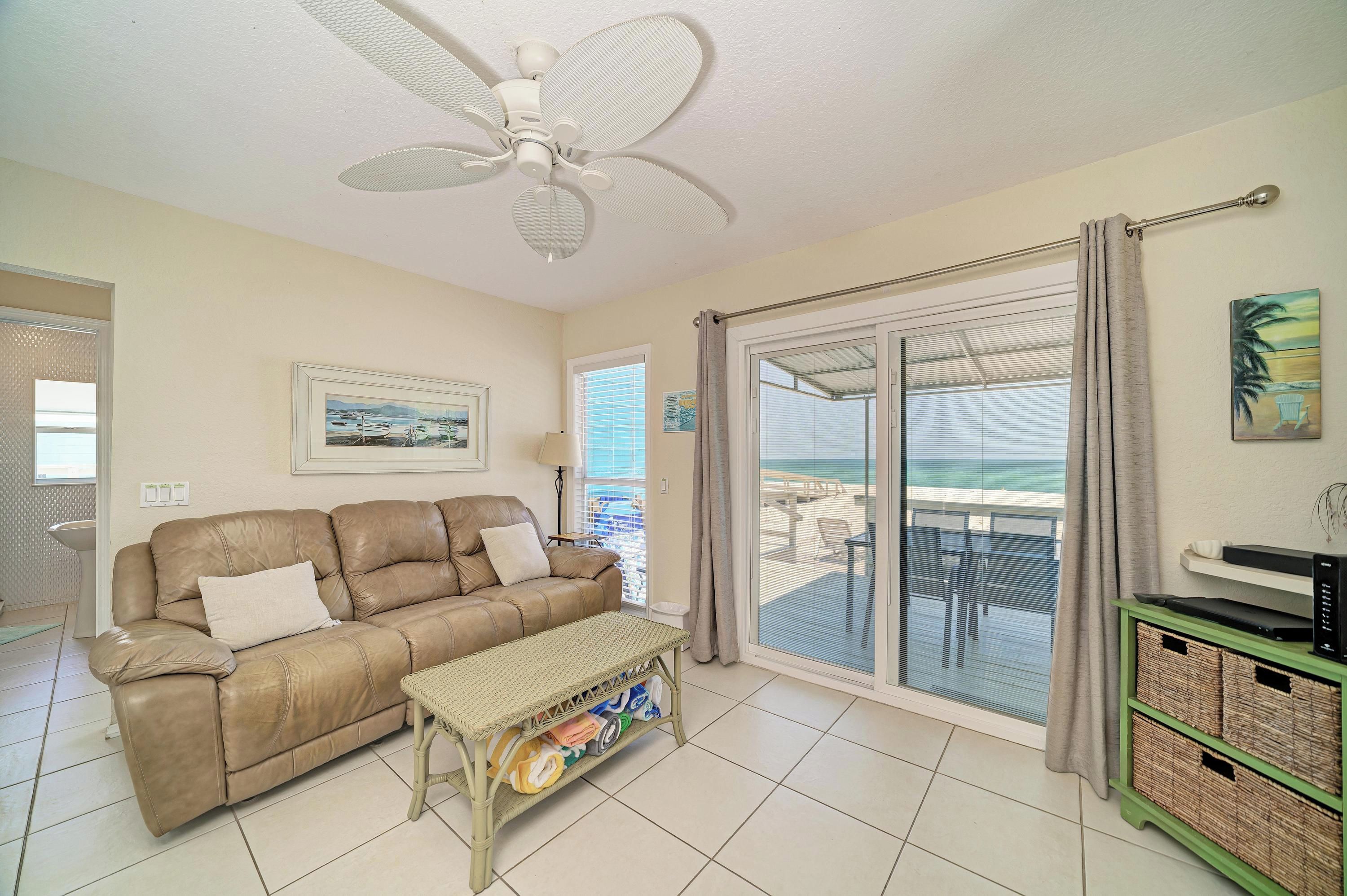 Sala de estar com sofá de couro bege, um banco trançado, ventilador de teto, porta de vidro deslizante que dá para uma varanda com vista para a praia e um quadro na parede.