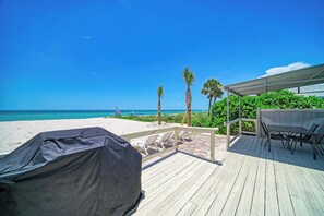 A wooden deck with a barbecue grill, outdoor seating, and a view of the beach and ocean.
