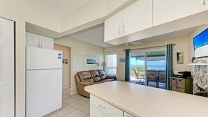 Ett kompakt kök med ett kylskåp, en matplats med en brun lädersoffa och utsikt över havet genom en skjutdörr i glas.