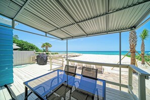 A covered outdoor seating area with a glass table and chairs, overlooking a beach with palm trees and the ocean.