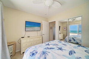 A bedroom with a bed, a dresser, a television on the wall, and a door leading to a balcony with a view of the ocean.