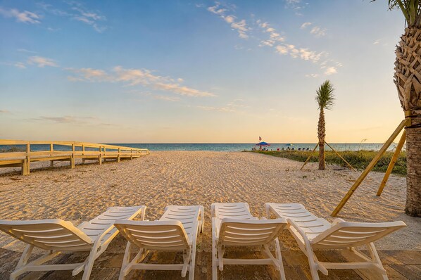 A beach with white lounge chairs arranged in rows facing the ocean, a wooden walkway, and a palm tree.
