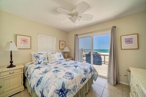 A bedroom with a bed, a nightstand, a lamp, a ceiling fan, a window with a view of the beach, and a door leading to an outdoor area.