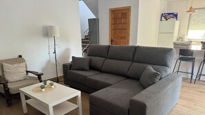 Un salon moderne avec un canapé d’angle gris, une table basse blanche agrémentée d’une plante, une chaise en bois, une lampe et un escalier menant à l’étage supérieur.