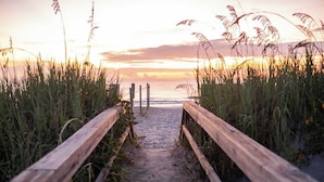 Ein hölzerner Weg führt zum Strand, gesäumt von hohem Gras.