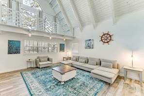 A spacious living room with a high ceiling, a large window, a wooden coffee table, and a sail wheel decoration.