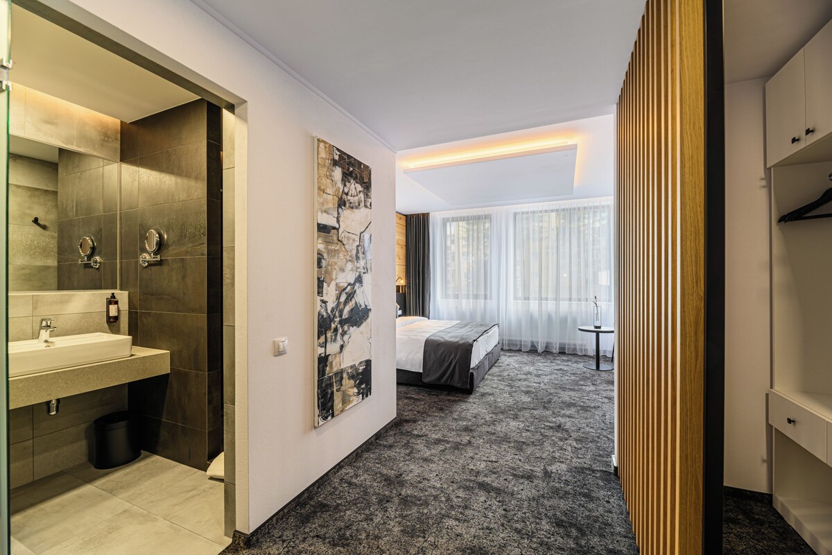 A hotel room with a bathroom, a bed, a bedside table, a window with curtains, and a wall-mounted artwork.