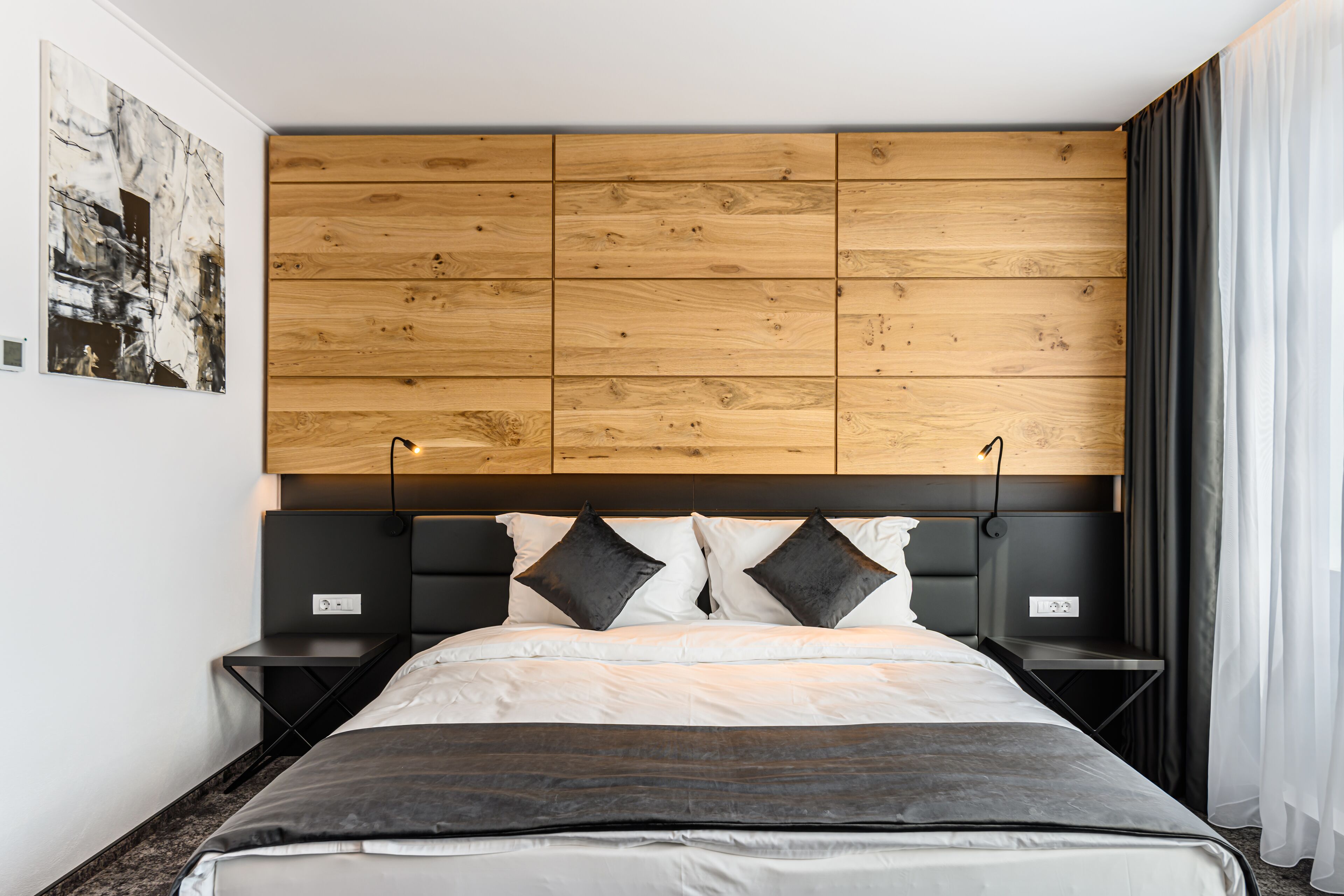 A modern bedroom with a wooden headboard, a bed with white and dark bedding, two nightstands, and a black lamp on each.