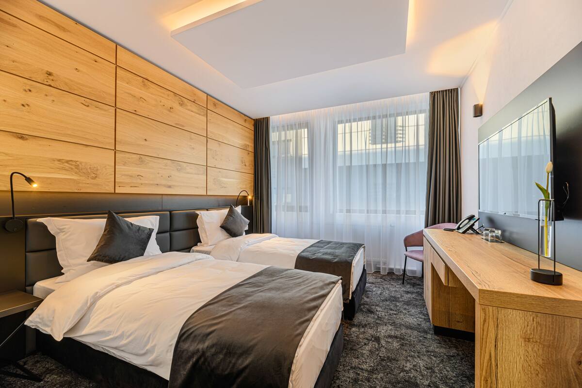 A hotel room with two beds, a wooden headboard, a desk, a chair, a mirror, and a large window with sheer curtains.