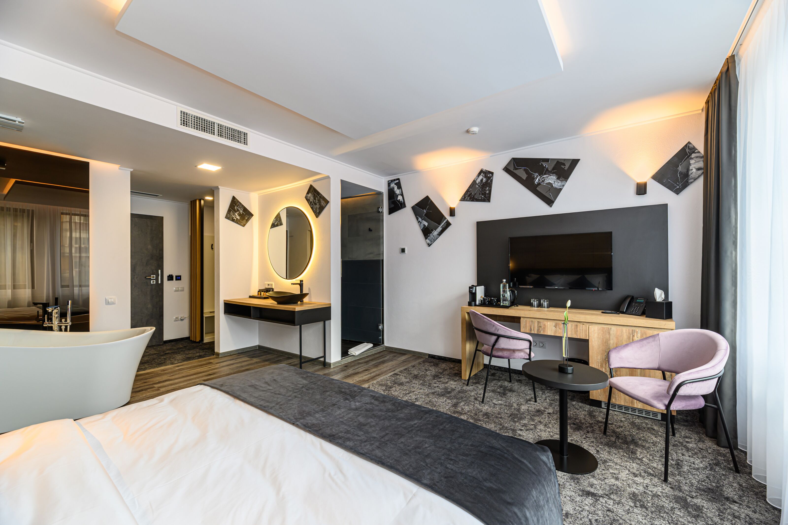 Une chambre d’hôtel moderne dotée d’un grand lit, d’un coin salon avec un bureau et une chaise, d’une coiffeuse avec un miroir et d’un téléviseur à écran plat fixé au mur.
