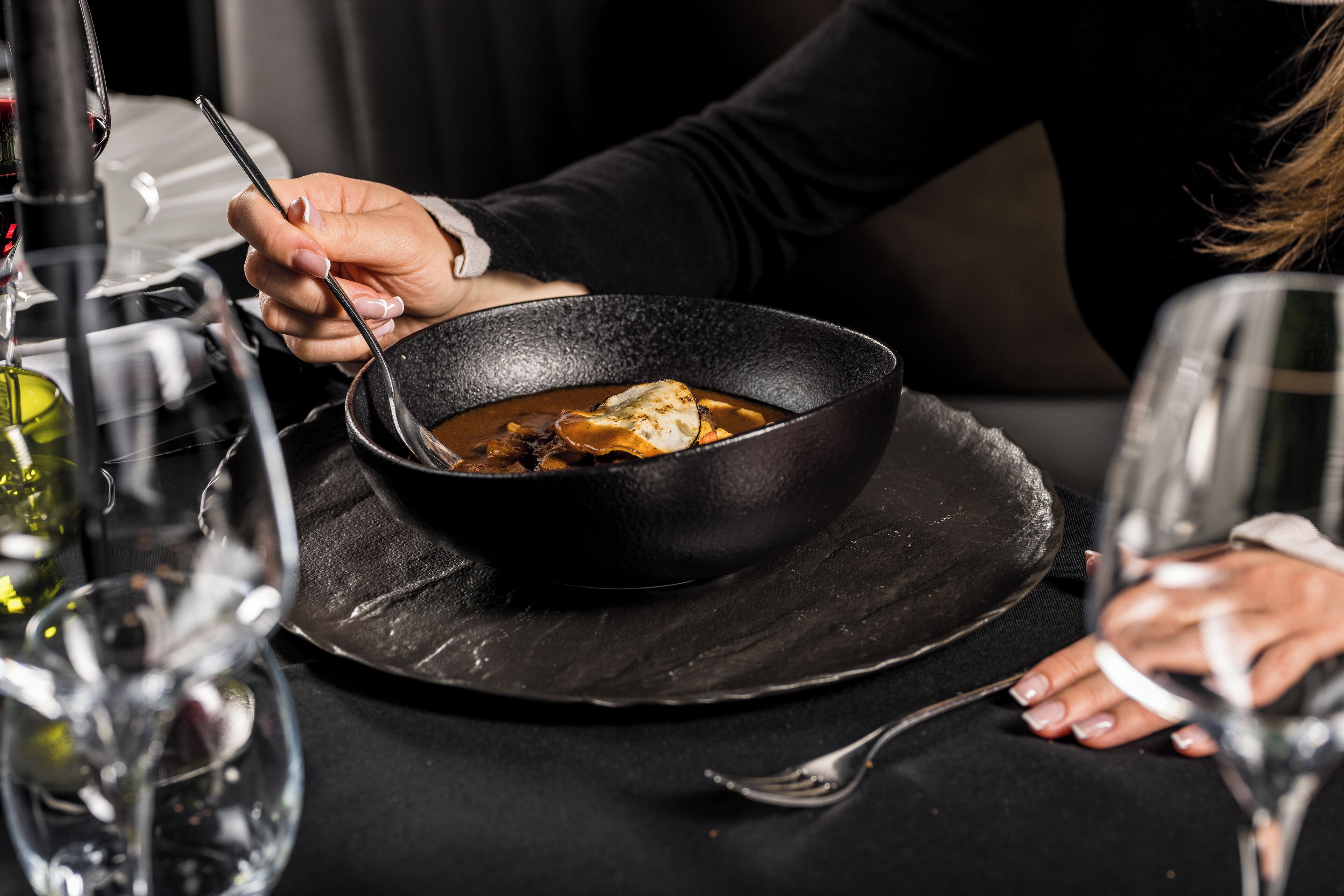 Una persona cenando en una mesa con un tazón de sopa, una copa de vino y un tenedor.