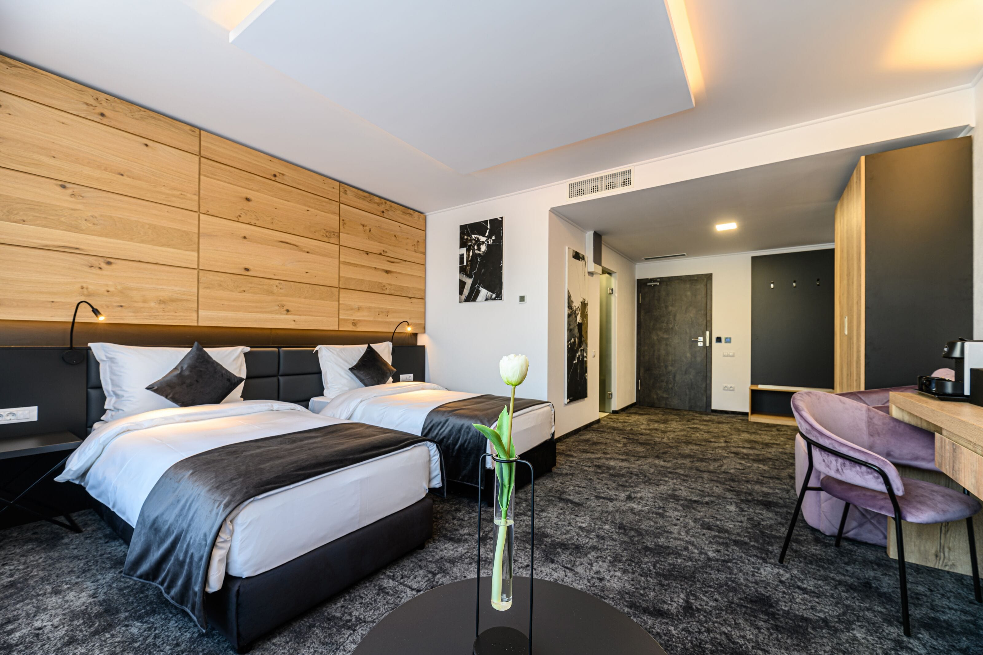 Une chambre d’hôtel moderne avec deux lits, un bureau et une chaise. Un vase avec une fleur est posé sur le bureau.