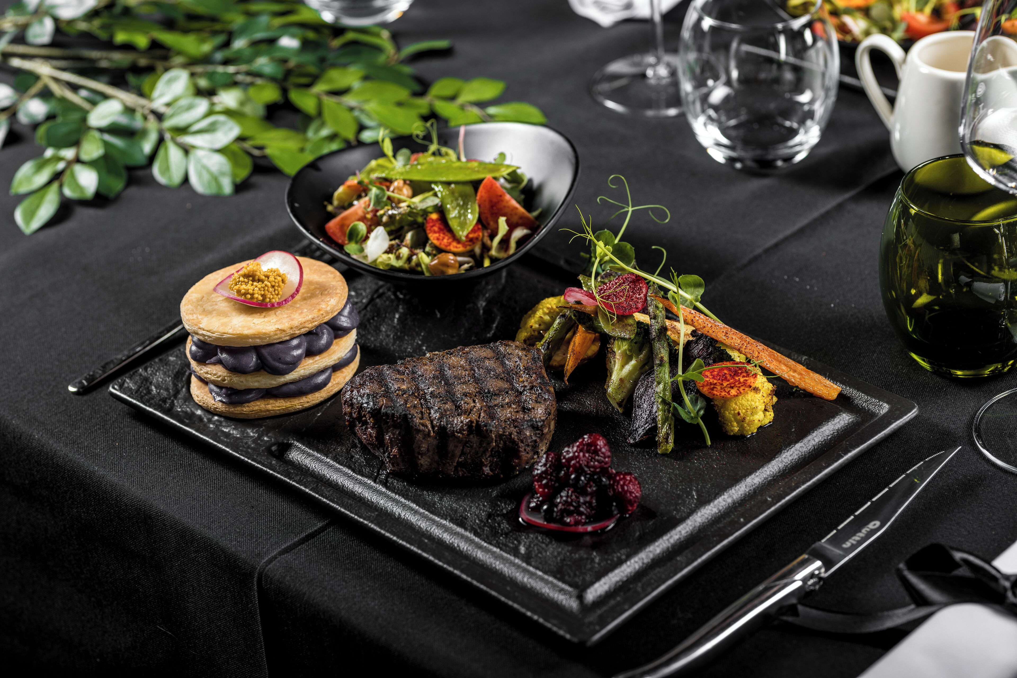 Et spisebord med sort dug, en steak, en dessert og en salat.