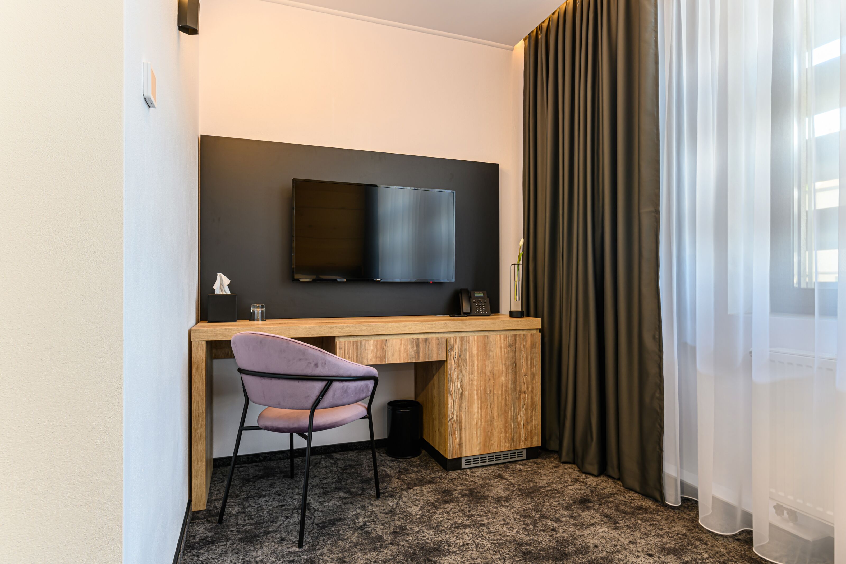 A hotel room with a wooden desk, a flat-screen TV, a purple chair, and a large window with curtains.