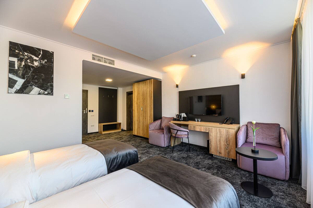 A modern hotel room with a bed, a desk, two chairs, a TV, and a painting on the wall.