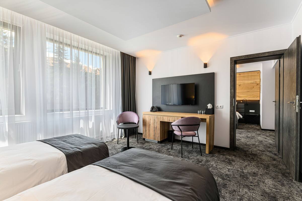 A hotel room with two beds, a desk with a chair, a TV, and a large window with sheer curtains.