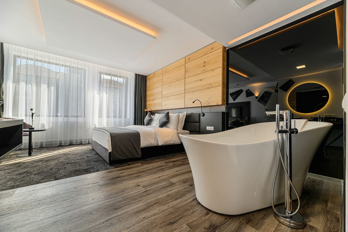 A modern hotel room with a large bathtub, a bed with a grey headboard, and a wooden panel wall.