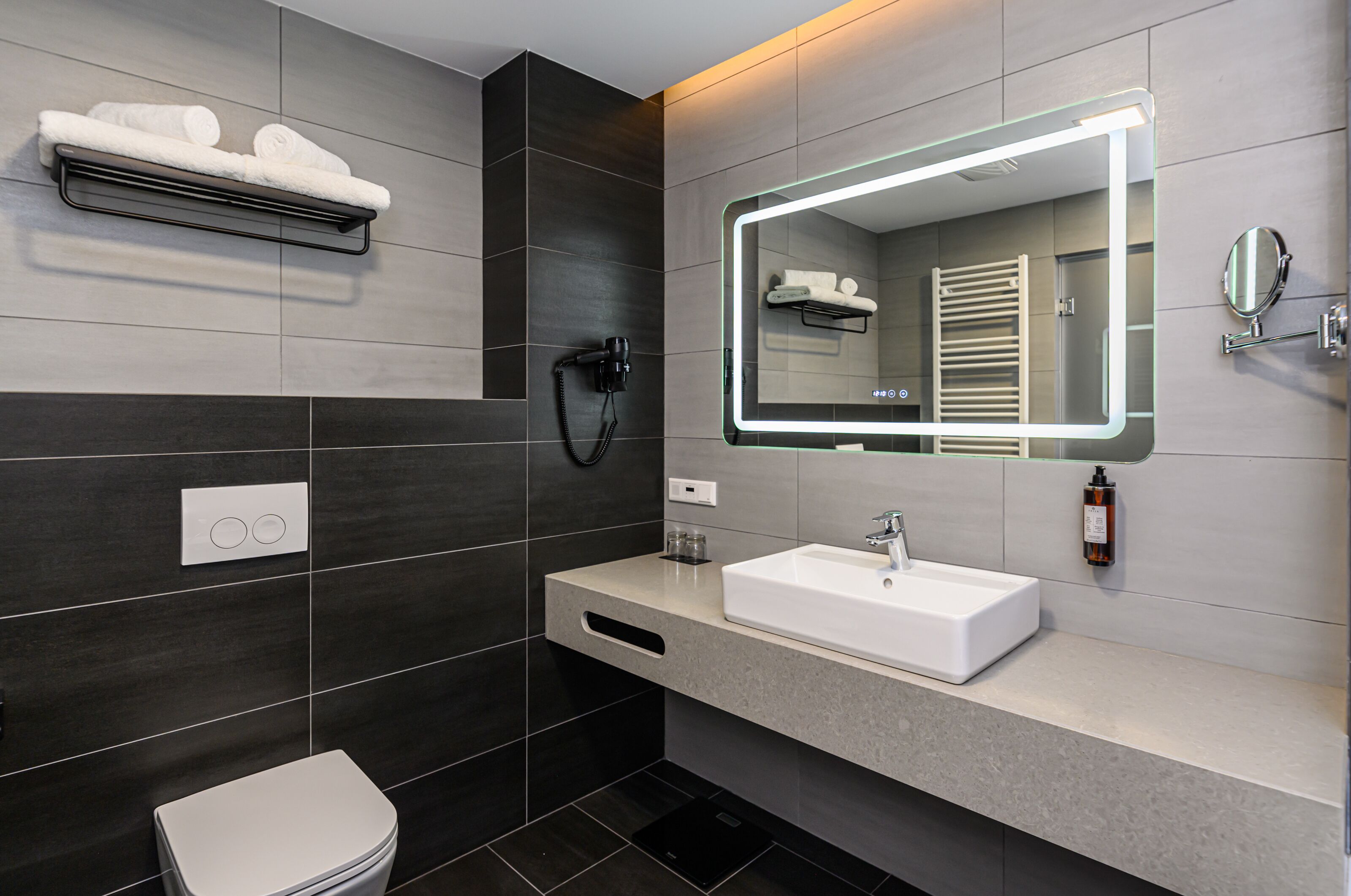 Un baño moderno con un lavamanos blanco, paredes de azulejos oscuros, un perchero, un secador de pelo y un espejo con iluminación integrada.