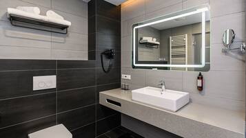 Un baño moderno con un lavamanos blanco, paredes de azulejos oscuros, un perchero, un secador de pelo y un espejo con iluminación integrada.