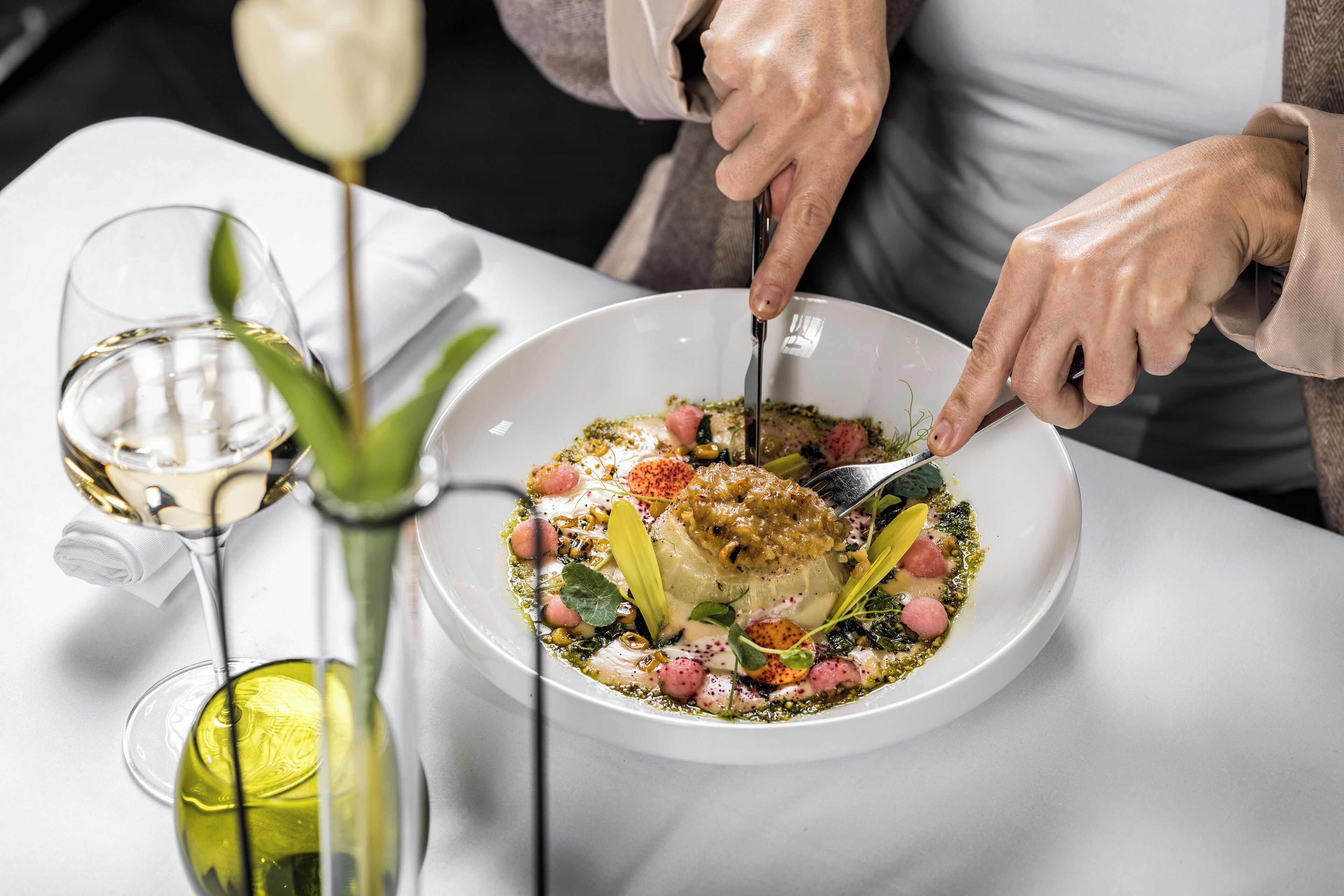 A person dining at a table with a white plate of food, a glass of white wine, and a green bottle.