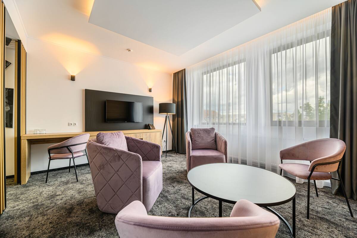 A modern hotel room with a flat-screen TV, a desk, and a seating area with cushioned chairs and a round table.