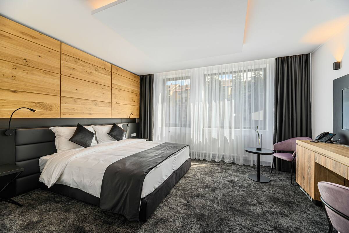A modern hotel room with a wooden headboard, a large bed with white and black bedding, a small round table, a desk with a chair, and a large window with sheer curtains.