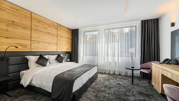 Una habitación de hotel moderna con cabecera de madera, una cama grande con ropa de cama blanca y negra, una mesita redonda, un escritorio con silla y un ventanal con cortinas translúcidas.