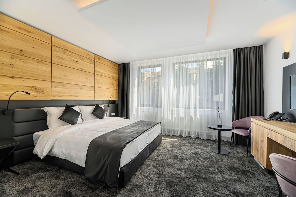 A modern hotel room with a wooden headboard, a large bed with white and black bedding, a small round table, a desk with a chair, and a large window with sheer curtains.