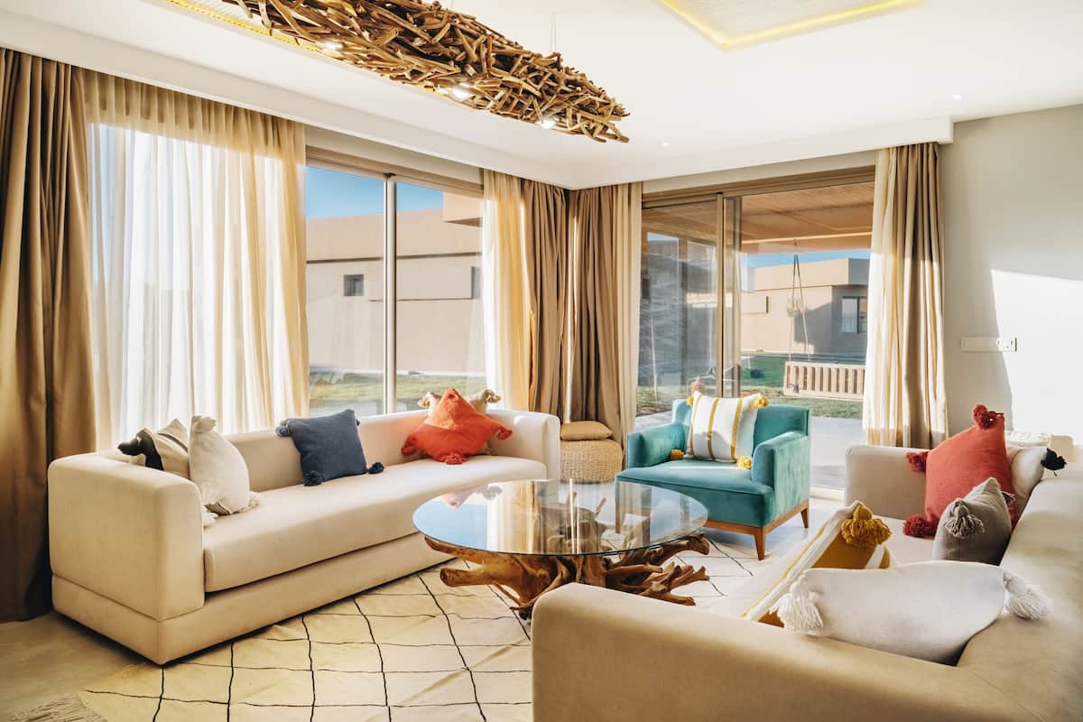 A modern living room with a glass coffee table, beige sofas, and teal armchairs.
