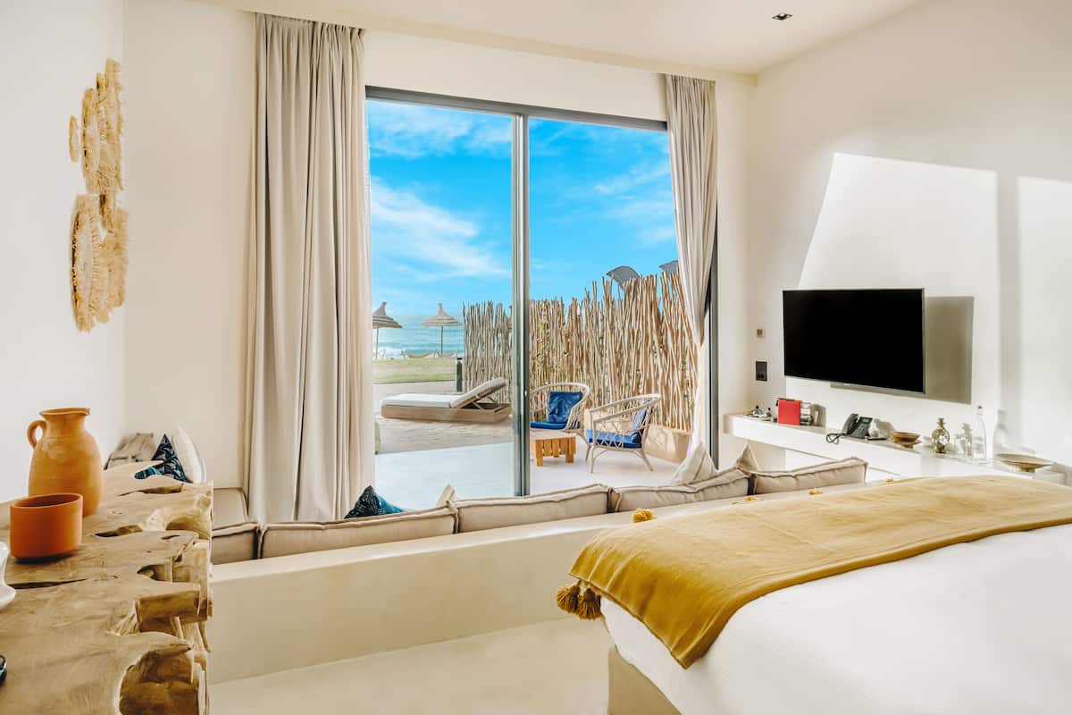A hotel room with a large window, a bed, a TV, and a seating area with a view of the beach.