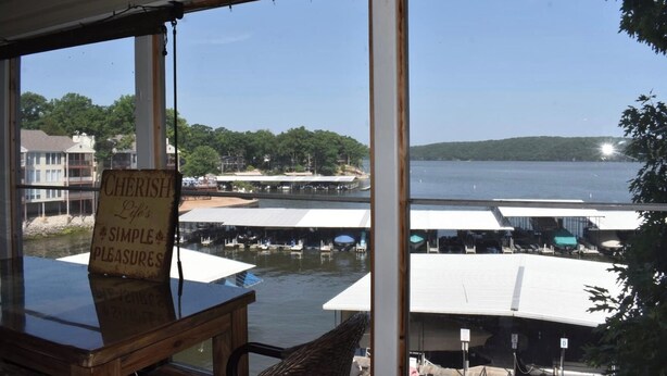 A view from a room with large windows overlooking a marina with boats and a dock area. A sign on the windows reads "CHERISH Life's SIMPLE PLEASURES." - Beautiful Lakefront Condo with boat slip, pool access and great location! (Osage Beach)