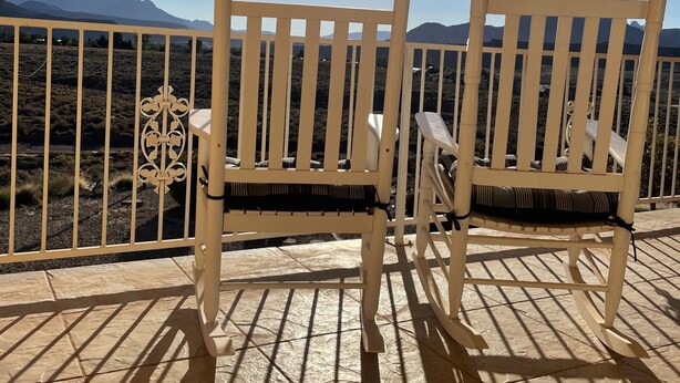 Zwei weiße Holzstühle mit schwarzen Kissen auf einem Balkon mit Blick auf Berge und einen klaren Himmel.