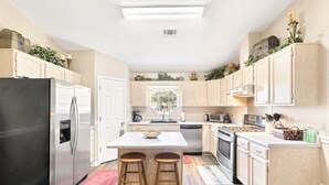 A modern kitchen with a stainless steel refrigerator, a white island with stools, wooden cabinets, and a window with a view. - Private Pensacola Getaway Minutes to Perdido Key! (Pensacola)