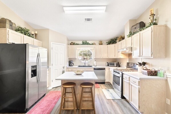 A modern kitchen with a stainless steel refrigerator, a white island with stools, wooden cabinets, and a window with a view.