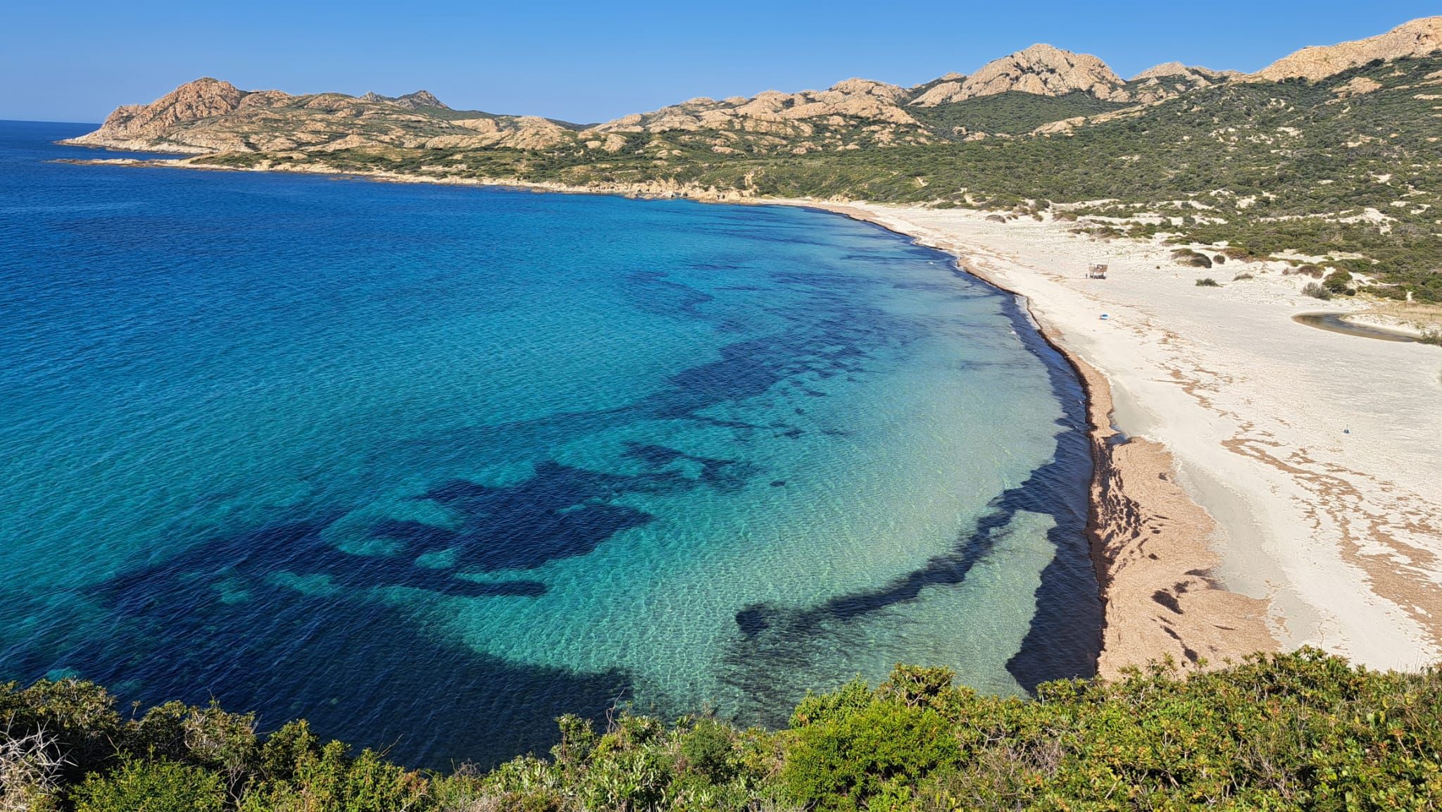 Un paysage côtier caractérisé par des eaux bleues et limpides, des plages de sable et, au loin, des collines rocheuses.