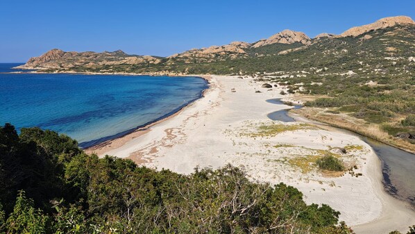 A coastal landscape with a sandy beach, clear blue sea, and rocky hills in the background.
