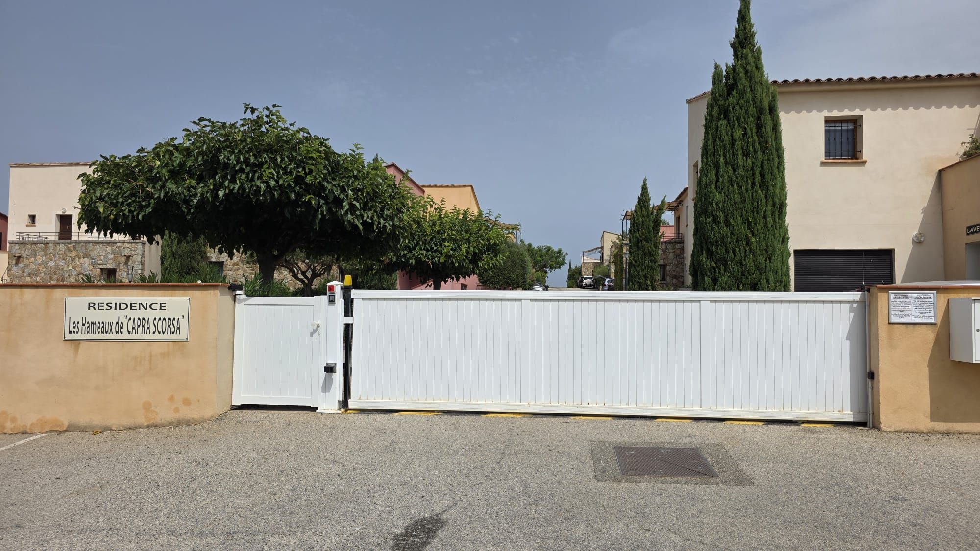 Une entrée fermée par un portail mène à une propriété appelée « Résidence Les Hameaux de CAPRA SCORSA », avec un portail blanc, un arbre et des bâtiments en arrière-plan.