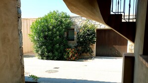 Vista desde una habitación con suelo de baldosas a un espacio exterior con un muro de piedra, un portón de madera y un arbusto grande con flores rojas.