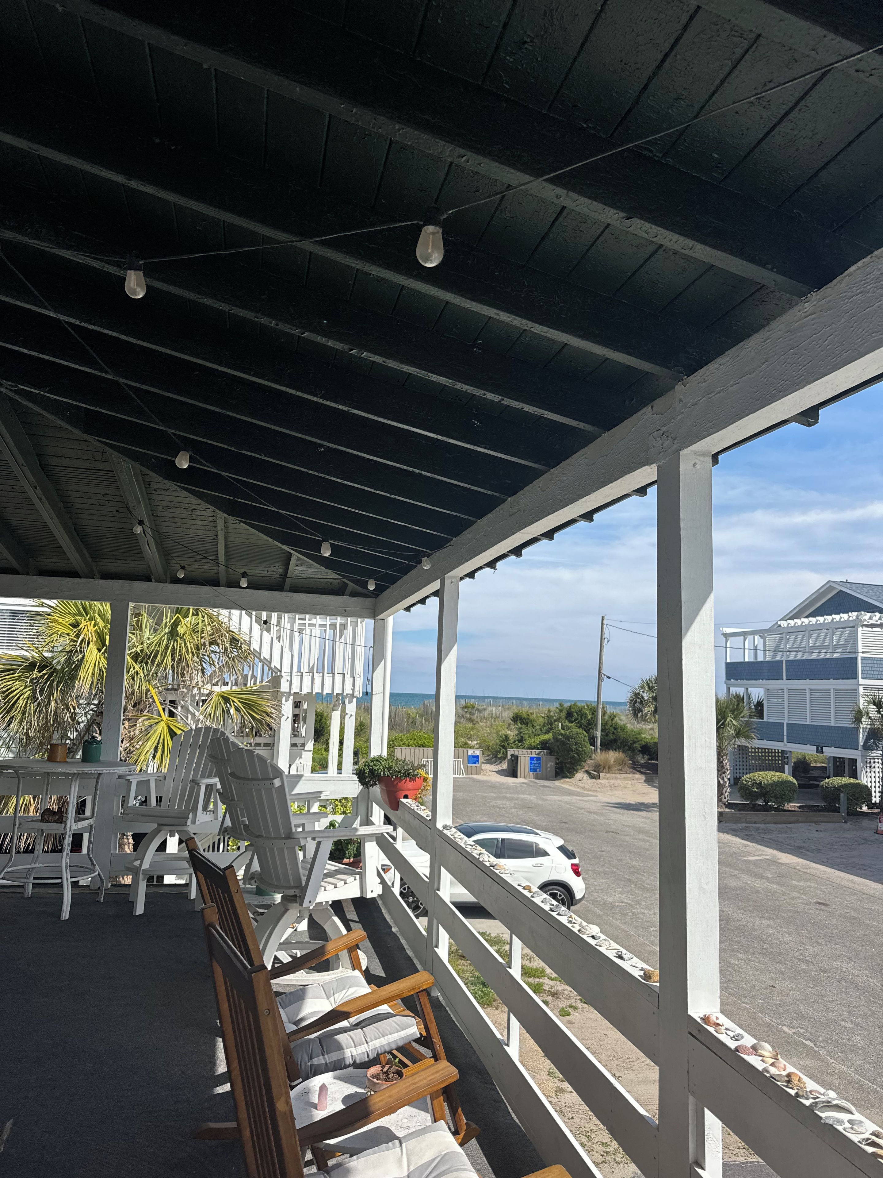 A covered outdoor seating area with chairs and tables, overlooking a beach and a parking area.