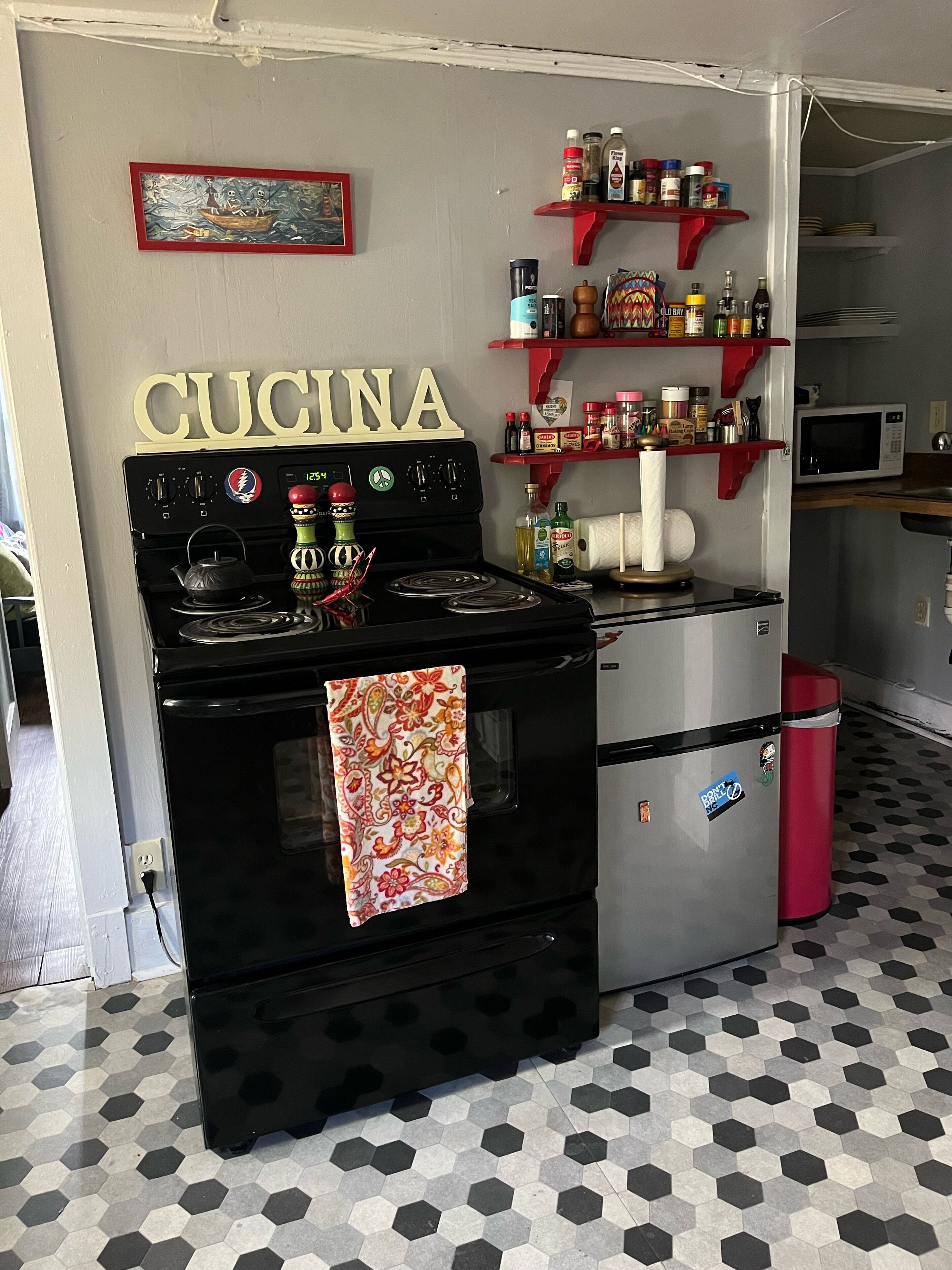 A kitchen with a black stove, a microwave, and a refrigerator. There are shelves with various kitchen items and a sign that reads 'CUCINA'.